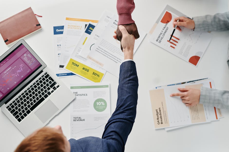 Business professionals engaging in a handshake over a desk filled with financial documents and a laptop.